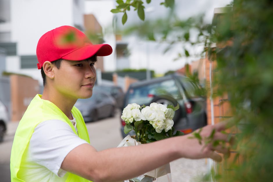 A young delivery person wearing a red cap and a neon yellow vest is holding a floral gift of white hydrangeas wrapped in white paper. They are reaching to hand over the bouquet to a recipient, with a