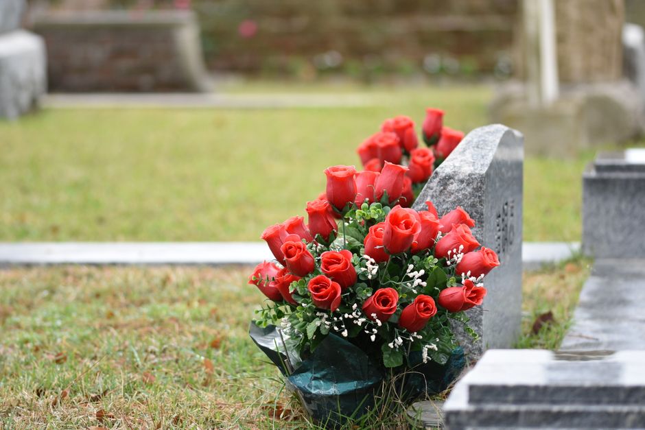 A bouquet of fresh, vibrant red roses with lush green foliage and delicate white baby's breath is placed at the base of a gray granite headstone in a cemetery setting. The roses are arranged in a trad