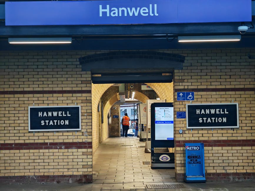 Entrance to Hanwell Station with beige brick walls and arched passageway leading to the platform area. Two black and white signs reading 'HANWELL STATION' are mounted on either side of the entrance. A