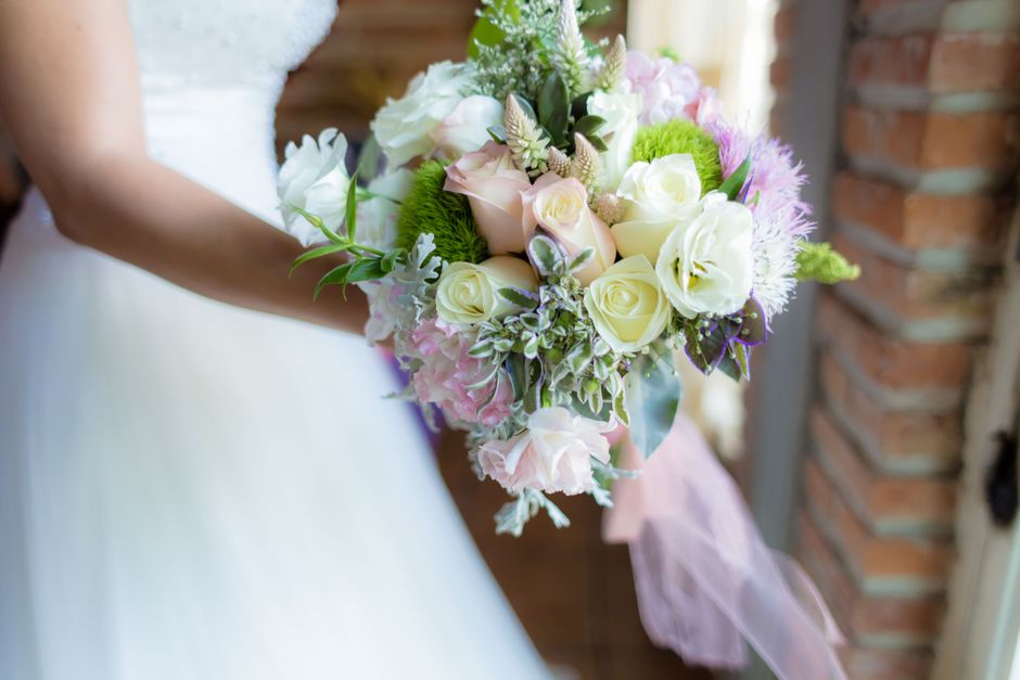 A person, dressed in a white wedding gown, holds a round bouquet arrangement of fresh flowers with both hands. The bouquet features white roses, pale pink roses, and cream-colored lisianthus, accented
