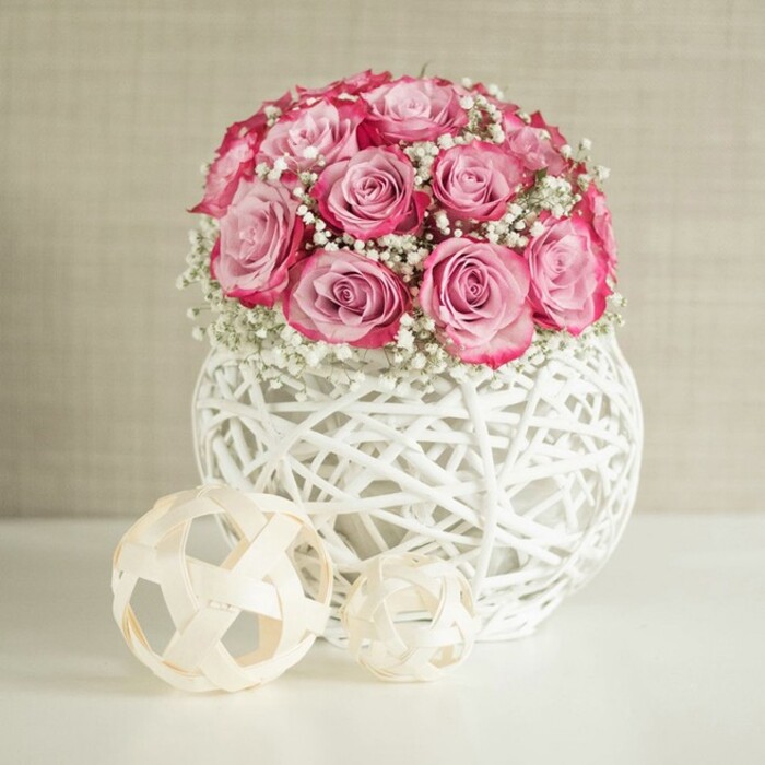 An elegant composition shows a rounded bouquet of pink roses and white baby's breath poised in a distinctive spherical vase, creating a serene, uplifting focal point that would sit beautifully in a living room overlooking Epping Forest. The roses form a dense, almost cushion-like dome, with each bloom at a slightly different stage of opening; some are still cupped, others fully unfurled, revealing layers of soft petals that shift from pale blush to a deeper, more saturated pink at their outer tips. This subtle ombré effect is enhanced by the contrast of the crisp white baby's breath threaded between the roses, their tiny clustered flowers creating a soft, misty texture that lightens the overall feel of the arrangement. The vase is crafted from white-painted natural fibres woven into an open, globe-shaped lattice, so the structure feels both airy and sculptural, with delicate shadows cast onto the pristine white surface beneath it. In front and slightly to the left, two decorative spheres made from thin, light wooden strips echo the vase's woven design; their warm beige tones and open framework add a gentle, organic balance to the cool white elements. The background is a muted, warm beige wall with a faint texture that adds depth without drawing attention away from the flowers, much like the understated interiors of many Epping homes and cafés tucked just off the High Street. Soft, even lighting bathes the entire scene, emphasising the tenderness of the pink tones and the craftsmanship of the arrangement. The mood is calm and sophisticated, making this design ideal for romantic gestures, thoughtful anniversaries, or simply adding a touch of everyday luxury to a hallway or bedroom.
