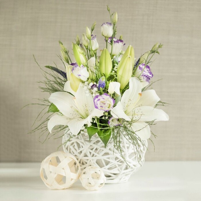 This image shows a sophisticated wedding table arrangement that feels both modern and romantic, as if styled for an intimate celebration in Epping. The centerpiece sits on a bright, white surface, its base a rounded ceramic-style vase in pure white, crafted with an open, basket-like weave that resembles finely worked rattan or wicker. Through the gaps in this lattice, you sense the stems and greenery inside, lending an airy, almost weightless quality. From this base rises a generous display of white Asiatic and oriental lilies; some trumpet-shaped blossoms are fully opened, their petals curving outward to reveal rust-colored stamens, while several sturdy, elongated buds remain tightly closed in fresh pale green, promising the arrangement will continue to evolve over the coming days. Woven among the lilies are numerous sprays of Eustoma, or lisianthus, in creamy white with delicately fringed petals tipped in lavender and soft violet. Some lisianthus blooms are open and ruffled, others gently unfurling, creating a layered, textural look reminiscent of bridal fabrics. Scattered throughout are tiny clouds of baby's breath, like soft white confetti, along with smaller deep purple accent flowers that provide subtle contrast without overwhelming the tranquil palette. The foliage is varied and abundant: feathery, fern-like greens create a halo of softness, while slightly broader leaves add structure and frame the flowers in rich, verdant tones. To one side, on the white tabletop, two decorative spheres made from light, woven natural material echo the vase's open-weave pattern, uniting the elements into a cohesive, design-led piece. The backdrop is a light beige wall with a subtle horizontal texture, evoking the calm interiors of Epping's homes and wedding venues, making this arrangement perfect for a reception table, remembrance service, or elegant dinner gathering.