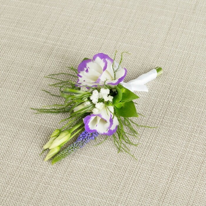 Set against a softly textured swathe of light beige linen, this boutonniere is captured in gentle, even lighting that lets its colours and textures unfold slowly to the eye. At its centre are two fully formed lisianthus flowers, each bloom a cup of creamy white petals edged in a deep, saturated purple, the ruffled margins almost ink-like where the colour is richest. They sit close together, like a pair in conversation, surrounded by tiny white filler blossoms that echo the look of baby's breath, giving the design a lace-like, ethereal softness. Rising just behind is a slim, upright stem of muscari, the small, bell-shaped florets crowded tightly in a column of dark grape-purple, adding height and a hint of woodland charm, as if it might have been snipped from a shaded corner of a garden near Epping Forest. Small, pale green buds nestle near the main blooms, the petals still folded in on themselves, hinting at freshness and the unfolding of the day ahead. A collar of greenery frames the flowers: fine, feathery fronds, likely asparagus fern, mingle with slightly broader, darker leaves, creating depth and a sense of movement, as though a breeze might ruffle them. The stems are drawn neatly together and wrapped in crisp white satin ribbon, the smooth band secured with a neat, understated bow, ready for easy pinning to a suit jacket. The arrangement feels timeless and refined, perfect for a groom, groomsman, or prom guest dressing for a celebration in Epping or the nearby countryside, capturing that blend of polished elegance and natural charm often requested by clients of Flowers Epping for their most meaningful occasions.