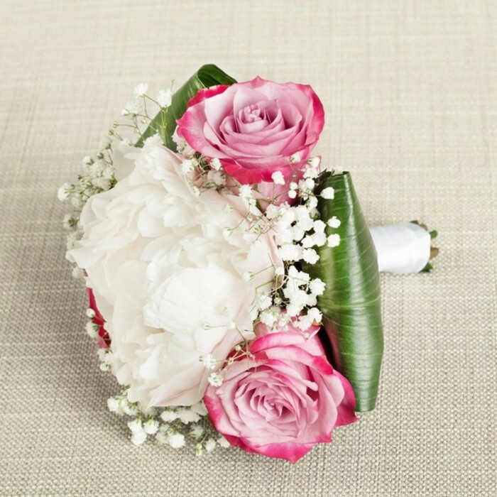 From a thoughtfully chosen angle, this exquisite bouquet appears as if carefully placed for a moment of pause on a softly woven, neutral fabric, echoing the understated elegance so loved in Epping's historic homes. Set amid a surrounding of lush, glossy green aspidistra or ti leaves, its heart boasts a luminous white peony-its petals soft, layered, and full, casting delicate shadows across their creamy expanse. Flanking this focal bloom, two vivid roses reveal petals edged in bold fuchsia gradually fading into blush pink and muted lavender, capturing the subtle transitions often witnessed in Epping's market gardens at dawn. Interspersed throughout, airy sprigs of baby's breath weave luminous punctuations, filling crevices and imparting a gentle, cloud-like softness. The bouquet is expertly finished with a gleaming white satin ribbon, binding the stems with elegance and care, ready to grace a wedding, anniversary, or local celebration. The soft, diffuse lighting illuminates each fold and curve, casting gentle highlights on the petals and enhancing the fresh vibrancy of each botanical element. Altogether, this arrangement reflects a timeless sophistication, local charm, and the dedication of Epping's skilled florists, making it the perfect floral gift or bridal accessory.