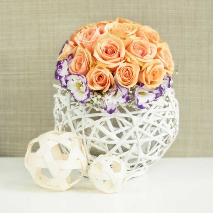 A meticulously crafted centerpiece rests on a gleaming white surface, its soothing palette and textures echoing the gentle charm of Epping's tree-lined streets. The arrangement is dominated by a perfectly rounded mound of roses in shades of soft peach, apricot, and salmon. Each rose is fully open and densely packed, their overlapping petals forming a smooth, domed silhouette that looks almost sculpted by hand. The blooms carry a sense of warmth and tenderness, reminiscent of early evening light filtering through the trees in Epping Forest. Just beneath this dome, a halo of white lisianthus unfurls, each blossom frilled and ruffled, with a striking border of deep violet-purple tracing the edge of every petal. The colour contrast between the warm roses and the cool purple trim is refined rather than bold, lending the piece a delicate, painterly quality. Interspersed between these lisianthus flowers are fine, cloud-like tufts of baby's breath. Their tiny white stars soften the transitions and create an airy, almost ethereal veil around the base of the roses. All of this floral detail is gathered into a rounded, bulbous container woven from white rattan or wicker. The open lattice design, with its crossing strands and visible gaps, casts gentle shadows on the surface beneath and gives a sense of lightness despite the arrangement's fullness. In front of the centerpiece, two decorative orbs made from pale, natural wood strands-one larger and one smaller-offer a subtle nod to rustic styling, tying in beautifully with local barn weddings and elegant receptions around Epping and nearby Loughton. A softly textured beige fabric backdrop completes the scene, its understated tone allowing the flowers to glow. The image conveys romance, composure, and artisan care, perfect for an intimate gathering or wedding breakfast in the Epping area.