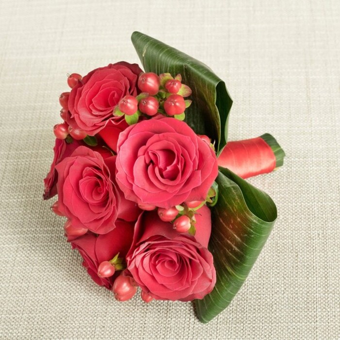 An exquisitely composed cluster of rich red roses and berries lies on a softly textured, light beige fabric, the warm neutral tones acting as a canvas for the bouquet's intense colour. Around six generous roses dominate the design, each bloom fully open yet still firm, with velvety petals spiralling from a tight central swirl into gently curved outer edges. The reds shift subtly from deep, wine-like crimson in the shadows to brighter scarlet where the soft light touches the upper petals, creating a sense of depth and movement. Between these roses, multiple sprays of small, round Hypericum berries sit like polished beads, their tones ranging from dark red to a softer, flushed pink. They are connected by slender green stems and tiny sepals that offer fine, almost delicate detail when viewed closely. On one side of the bouquet, a pair of broad, dark green leaves, possibly aspidistra, have been expertly shaped and wrapped partway around the flowers, forming a structural frame that both supports and accentuates the arrangement. Their glossy surface and clear veining contrast beautifully with the matte fabric beneath. Around the stems, a deep red satin ribbon is neatly wrapped and secured, the sheen of the ribbon mirroring the richness of the petals. The lighting is even and gentle, free of harsh glare, so the bouquet appears serene and carefully staged, as if ready to be lifted and handed to a loved one. This design, ideal for anniversaries, Valentine's gestures, or a romantic dinner in Epping-perhaps near Epping Forest or in a cosy home off Theydon Road-embodies classic passion infused with the care of a skilled, local florist.
