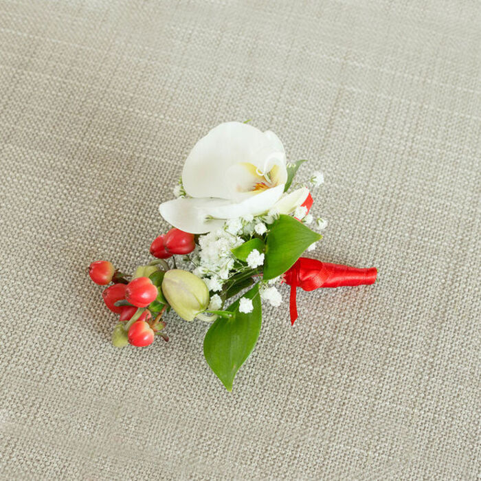 An elegant boutonniere is captured from a gently elevated viewpoint, placed against a subtly textured, light beige woven fabric that recalls natural linen often seen in rustic venues around Epping. At its center is a pristine white Phalaenopsis orchid bloom, the petals broad, smooth, and slightly curved, giving the flower a serene, sculptural quality. The orchid's throat reveals a delicate blend of creamy yellow and soft orange, adding just enough warmth to draw attention to the intricate heart of the blossom. Surrounding this focal flower, fine clusters of white baby's breath create a halo of tiny, star-like blooms, their airy structure softening the bold presence of the orchid and adding a sense of lightness and movement. Threaded through and just below, a concentrated cluster of rounded red berries glows with a polished, almost lacquered shine, bringing a joyful, festive note reminiscent of winter celebrations or Christmas parties held in the town's cosy pubs and event spaces. Interspersed among the berries are a few lighter green buds, which introduce a subtle variation in tone and hint at newer growth. Lush green leaves in different sizes and shapes-some broad and ovate, others slimmer and more elongated-frame the arrangement, adding depth, natural contrast, and a freshly picked feel. All the stems are cleanly gathered and bound with a bright red satin ribbon, wrapped tightly and finished in a neat, compact knot with short, elegant tails. The overall composition suggests careful craftsmanship and thoughtful design, making this boutonniere a refined choice for grooms, groomsmen, or formal guests attending weddings, proms, or special occasions in and around Epping. Its combination of crisp white, vivid red, and natural greenery creates a timeless, celebratory look that feels both classic and distinctly local.