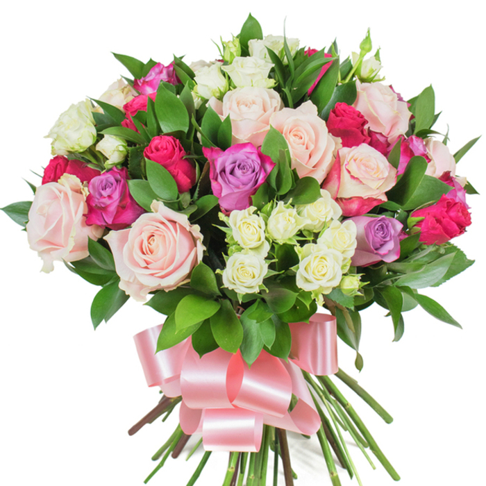 A dense, rounded bouquet of mixed roses and rich greenery is presented against a stark white background that highlights every petal and leaf. The composition favours large, traditional roses in soft blush-pink and creamy white, some opening wide to reveal satiny inner petals, others poised as elegant buds. Scattered among them are stronger magenta or hot-pink roses that animate the palette with joyful brightness, and a few lavender roses add soft, unexpected coolness to the mix. Tiny spray roses in pale cream provide delicate clusters that break the larger shapes into textured layers, lending fullness and a handcrafted feel. Deep green, glossy foliage-likely ruscus or salal-threads through the bouquet, its elongated leaves creating a natural framework that accentuates the blooms' colour. The stems are gathered securely and wrapped with a broad, baby-pink satin ribbon tied into a prominent bow whose smooth sheen catches studio light and complements the bouquet's romantic tones. Close to the flowers one can imagine a faint, classic rose scent with a hint of green leafiness; together the tactile softness of petals and the sleekness of the ribbon make the piece feel lovingly assembled. As a florist familiar with Epping's community, I picture this bouquet chosen for birthdays, anniversaries, or to celebrate someone special after a walk near St. John's or a weekend browse of the High Street market - a gift that reads as both joyful and elegantly sincere.