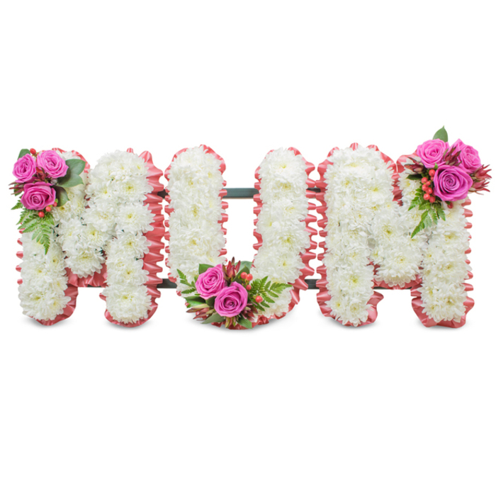 A poignant, expertly arranged memorial piece spelling MUM in large, connected letters, shown against a simple white backdrop to emphasise its handcrafted detail. Each letter is formed from thick clusters of immaculate white chrysanthemums, their compact heads lending a plush, tactile surface that reads as serene and pure. A soft, ruffled trim of rose-pink fabric outlines each letter, introducing a gentle motherly warmth that complements the white. Between the letters a dark green support frame is subtly visible, the practical armature that keeps this standing tribute steady during a funeral service or at a graveside. Three artfully placed posies enliven the design: the first M features a small bouquet of deep fuchsia roses, dark glossy foliage and tiny coral hypericum berries at its upper left; the U is balanced with a matching cluster at the base of its curve; and the final M mirrors these accents at its upper right. The interplay of textures - crisp chrysanthemum faces, velvet rose petals, shiny berries and feathery fern - offers both visual and tactile comfort, and a delicate floral scent drifts from the roses. I picture this piece in a quiet Epping chapel or set beside a family grave near Bell Common, where it communicates love, respect and a gentle farewell. The arrangement reads as a personal expression of remembrance, suitable for a memorial service, wake or graveside dedication for a much-loved mother.