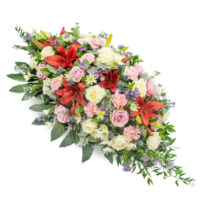 Set against a clean white backdrop, this elongated casket spray appears almost to float, its abundance of flowers and greenery creating a soft, flowing silhouette from a wider crown at the upper left to a slender point at the lower right. Deep red lilies, likely stargazer or Oriental varieties, rise proudly from the centre and through the length of the arrangement, their star-shaped blooms with curved petals offering a bold, heartfelt statement of love and respect. Surrounding these dramatic flowers are creamy white, ivory, and blush pink roses, some in tight buds, others fully open with layered, velvety petals that suggest tenderness and grace. Light pink carnations, full and ruffled, knit the larger blooms together, while clusters of small white chrysanthemum or daisy-type flowers scatter gentle highlights throughout the spray. Threaded subtly between are steel-blue thistles and tiny lavender-blue filler flowers, perhaps statice or veronica, which introduce cool, misty tones reminiscent of an early morning walk through Epping Forest. The colour harmony of reds, pinks, whites, and purples feels considered and calm rather than loud, lending a quiet dignity to the design. Beneath and beyond the flowers, lush green foliage spreads out in different shapes and textures, from broad, glossy leaves to lighter, feathery fronds that soften the outline and add a sense of movement. This is clearly a tribute crafted for a moment of farewell-a casket spray that would sit beautifully in a local Epping chapel or crematorium, offering comfort to family and friends as they gather to remember a loved one with beauty, colour, and a final, graceful gesture.