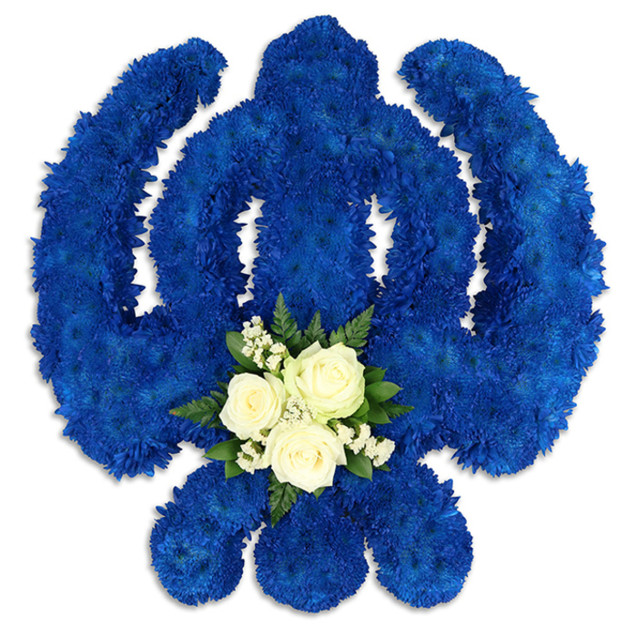 This photograph shows a beautifully composed Sikh Khanda floral tribute, presented on a clean white background to emphasise its bold outline and rich colours. The entire Khanda emblem - the powerful central sword, the perfectly round chakkar, and the two sweeping, symmetrical kirpans - is crafted from hundreds of small, tightly clustered royal blue blooms, likely chrysanthemum florets. Each head nestles into the next, creating a thick, cushion-like layer of petals that gives the piece a plush, almost three-dimensional feel. The blue is deep and saturated, reminiscent of the cool, calm atmosphere on an early morning walk through Epping town centre, and it conveys strength, dignity, and spiritual depth. At the base of the central sword, just where the eye naturally comes to rest, sits a focal posy of fresh white flowers. Three immaculate white roses are arranged close together, their petals spiralling softly outward, surrounded by fine sprays of delicate white filler flowers that look like baby's breath, and edged with short, glossy dark green leaves. This smaller arrangement provides a gentle contrast to the surrounding blue, adding a note of peace, comfort, and human tenderness to the strong religious symbolism. Soft, even lighting highlights the texture of each petal and leaf, while faint shadows underneath hint that the tribute is slightly raised, suggesting presence and importance. It is the kind of thoughtful, hand-crafted Khanda tribute that might be placed at a Sikh funeral or remembrance service for a family from Epping, offering a visually powerful yet serene expression of faith, remembrance, and enduring love.