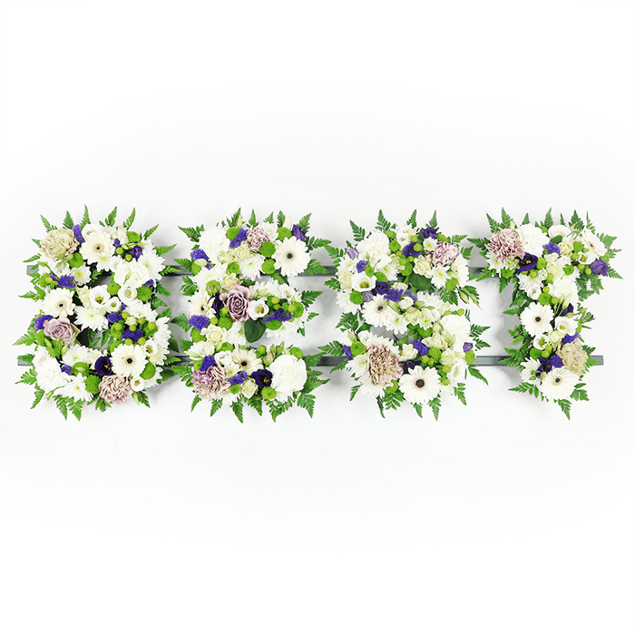 A horizontal, handcrafted floral lettering piece spells out the uppercase word BEST in a serene palette of white, mauve and green, photographed on a pristine white surface to emphasise each intricate detail. The letters B, E, S and T are each densely composed of large, pure white gerbera daisies-their dark centres providing quiet contrast-alongside soft white lisianthus and full, creamy carnations that convey peace and remembrance. Interspersed are refined touches of soft lavender and mauve roses and deeper violet filler blooms, which lend an air of dignity and warmth without overwhelming the gentle whites. Throughout the letters, compact green button chrysanthemums give small pops of vibrant freshness and textural variety, while feathery fern fronds trace the outer edges, creating graceful, natural borders that soften the overall silhouette and lend balance. The flowers are anchored on a dark structural base that is subtly visible in places, its neutral tone helping the blossom colours read clearly under even, diffused lighting. Together the layered textures-from the silky rose petals to the rounded gerbera faces and the airy fronds-create a composition that reads as both elegant and heartfelt. The arrangement's mood is contemplative and tender, ideal for use as a sympathy tribute at wakes, memorial services or cremations in the Epping area, perhaps displayed near a chapel on the High Street or at a local community centre. It's a personalised floral message that speaks of deep esteem and lasting memory, crafted with the considered hand of a florist who understands how to honour a life through flowers.