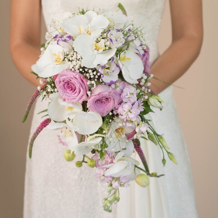 Crafted with sensitivity and expertise by a florist attuned to Epping's traditions, this photograph offers a close, evocative perspective of a bride holding a lush waterfall bouquet-the central focus against a softly blurred, neutral background. The bride's white gown, featuring subtle lacework that catches muted light, signals grace and poise as she gently clasps the fragrant arrangement. Dominating this composition are immaculate white Phalaenopsis orchids, their delicate petals and golden centers radiating quiet luxury, reminiscent of floral displays along Epping High Road. Intertwined are pink and lavender roses-some open to their full, romantic potential, others tightly furled, speaking to new beginnings and tender promises. Baby's breath is woven throughout, its airy form dissolving sharp edges and highlighting the bouquet's fluid, organic drape. Smaller purple accents, perhaps sweet pea or lavender stock, appear like delicate brushstrokes, adding both color and a gentle, local touch. The bouquet's cascade is enhanced by striking fuchsia amaranthus that trails with velvet-like texture, balancing structure and whimsy. Hints of tender green foliage and tiny unopened buds nod to the fresh beauty found in Epping's early summer landscapes. With every bloom carefully considered, the image not only celebrates bridal beauty but represents a local love story-designed for romantic ceremonies in Epping's charming environs and offering inspiration to those seeking floral artistry with both classic and modern resonance.