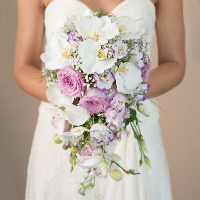 A striking, vertical composition showcases a bride in Epping tenderly supporting a magnificent, cascading wedding bouquet, the scene suffused with the soft light and tranquil atmosphere characteristic of local civil ceremonies. The bride, seen from chest to waist, is clad in a classic white strapless gown adorned with elegant lace patterns that add a layer of artistry to the image's lower frame. The bouquet, designed in an elegant tear-drop style, overflows with a harmonious mix of creamy whites, blush pinks, and gentle lavender hues. Prominently displayed are opulent white phalaenopsis orchids, their luminous, wide petals and golden centres exuding luxury and freshness-much like those selected for celebrations around Epping's historic market square. Nestled between the orchids are romantic garden roses in balmy blush and soft pink, their velvety layers reminiscent of petals freshly gathered after a gentle rain in local gardens. Delicate lavender stock and light purple lisianthus, some twinned with dark purple edges, lend playful texture and subtle depth while airy sprays of white gypsophila drift throughout, echoing the lightness of a morning breeze over Stonards Hill. Water-fresh green foliage and slender stems complete the natural cascading shape, creating a dynamic, organic flow. The bouquet's serene palette and lush textures are a testament to local Epping floristry expertise-a perfect choice for couples seeking sophistication and romance set against the backdrop of this picturesque Essex area.
