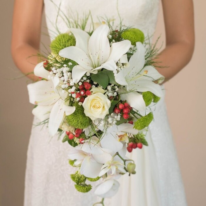 In this detailed portrait, a bride is pictured grasping an opulent cascading bouquet-a testament to the refined floristry found at Flowers Epping. The bouquet's artistry is anchored by a cluster of majestic, pure white lilies, their trumpet-shaped petals elegantly spattered with fine copper markings and bringing a dramatic flourish admired by many at Epping's historic wedding venues. The arrangement's heart lies in a delicately swirled, cream-coloured rose, its subtle fragrance mingling with memories of nearby wild rose gardens. Interwoven among these classic blooms are airy sprays of gypsophila, imparting a diaphanous texture, while bold, spherical, lime-green cushion chrysanthemums inject a flash of contemporary style evocative of springtime walks through Epping Open Space. Accents of bright, round red Hypericum berries punctuate the display, their vibrant color reminiscent of the festive market days in the town centre. The iconic teardrop silhouette is formed by a veil of pristine white Phalaenopsis orchids, cascading elegantly below the main cluster and adding both length and fluid grace to the design. Fine blades of grass and select leafy greens curve around the bouquet, framing it against the bride's softly textured, ivory lace gown, which glows tenderly in diffused daylight. The background remains gently out of focus, ensuring attention lingers on the unique interplay of colors, forms, and textures. This luxurious, thoughtfully composed bouquet serves as both a focal point and a subtle tribute to Epping's natural beauty, ideal for brides seeking an unforgettable blend of classic opulence and fresh appeal.
