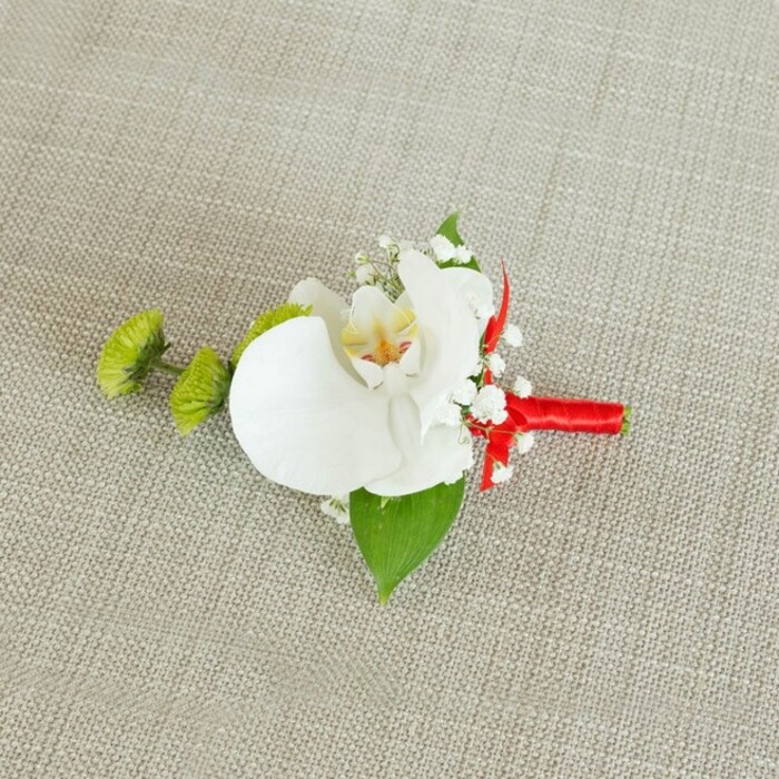 Displayed against a pale beige linen-style fabric, this sophisticated boutonniere is captured in soft, even lighting that gently emphasises every petal, leaf, and thread. The neutral background, with its faintly visible weave and natural, organic feel, creates a calm stage for the floral details. At the centre sits a flawless white Phalaenopsis orchid, its large, gracefully arched petals opening outward like calm wings. The orchid's inner throat reveals an intricate blend of soft yellow and subtle reddish-orange veining, a fine detail that breaks the purity of the white in a way that feels both refined and inviting. Encircling the orchid, clusters of delicate baby's breath form a light, downy frame of tiny white blooms, adding movement and a hint of rustic charm, much like the wildflowers you might notice along quieter lanes leading out of Epping toward the countryside. To the left, two rounded lime-green chrysanthemum buds, tightly packed with tiny, spiky petals, stand out as bright accents. Their fresh green shade and playful shape introduce a modern twist, injecting vitality without stealing focus from the orchid. Beneath the flowers, a broad, deep green leaf acts as a natural base, anchoring the design, while a smaller, slightly darker leaf near the stem adds layered depth. The stems are neatly wrapped in a bold crimson red satin ribbon, its smooth sheen contrasting beautifully with the rougher linen texture below. The ribbon is wound firmly and finished in a subtle bow or knot, lending a polished, celebratory touch. This carefully crafted piece feels ideal for a wedding party in Epping, perhaps for a groom making his way from home to a nearby ceremony, wearing a floral detail that combines classic romance with a fresh, contemporary edge.