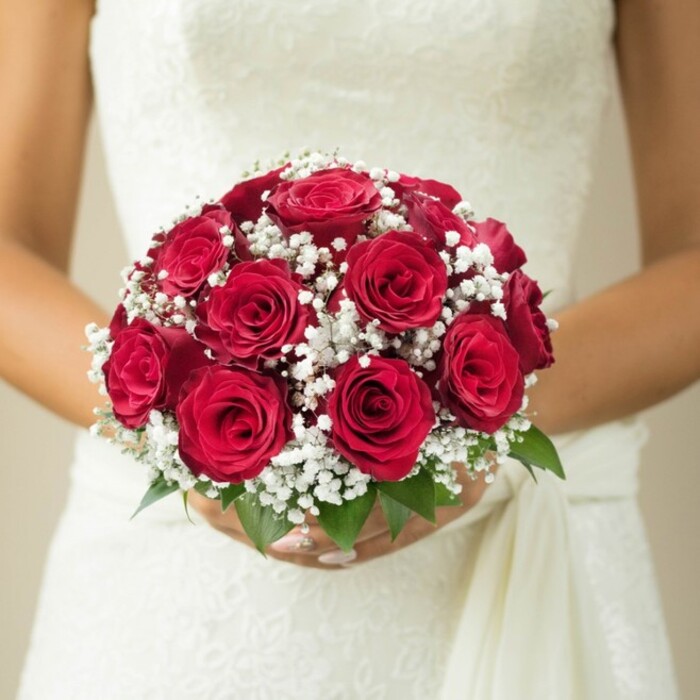 This deeply evocative photograph, set against the tranquil ambiance of Epping, captures a bride holding a vibrant, domed bridal bouquet that epitomizes classic wedding elegance. Dominating the composition are striking, full-bodied red roses, their petals a tapestry of velvety richness-some blooms tightly furled, others more open, each suggesting a different beat in the rhythm of romance. Weaving through these passionate blooms are generous clusters of white gypsophila, or baby's breath, whose cloud-like sprigs add an airy, textural softness and create a striking counterpoint to the deep reds. Below, fresh green leaves extend slightly, accenting the bouquet with a hint of the natural beauty that surrounds Epping's High Street and open spaces. The bouquet is tenderly cupped in the bride's manicured hands, while behind the flowers, her wedding gown reveals delicate threads of lace or embroidery, a subtle nod to time-honoured bridal styles. A gentle sash or trailing fabric drape falls along her side, and the light, creamy backdrop is softly blurred to frame this moment of anticipation and joy. Designed with the care and attention to detail known among Epping's best florists, this bouquet is a loving celebration of tradition, modern artistry, and the singular romance only a wedding in Epping can inspire.