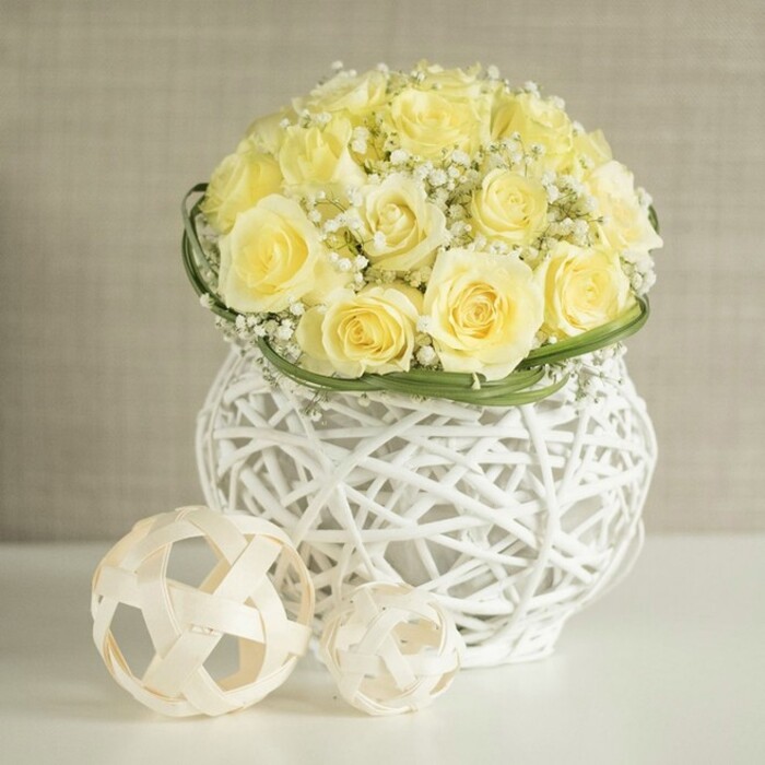 A chic, contemporary centerpiece sits poised on a flawless white surface, ideal for a wedding table in an Epping barn venue or a modern home near Bell Common. At its heart is a tightly composed dome of pale yellow roses, each blossom at its fullest, with petals unfolding in generous spirals that suggest softness to the touch and a gentle, powdery fragrance. The roses are arranged so closely that they appear to form one continuous mound of creamy colour, yet you can still appreciate the individuality of each bloom. Interspersed throughout, fine threads of white baby's breath, or gypsophila, emerge like tiny stars scattered between clouds, bringing both texture and airiness. A polished band of bright green foliage hugs the base of the bouquet, forming a clean ring that visually lifts the flowers and neatly defines their outline. This lush cluster is cradled in a white woven vase with an open lattice structure, reminiscent of handwoven rattan or wicker, its rounded silhouette gently pinched at the centre to give an elegant, hourglass feel. Light filters through the woven strands, casting delicate shadows that dance across the tabletop. In the foreground, two decorative, spherical accents made from slender, natural-coloured strips of wood or rattan rest casually, one larger, one smaller, echoing the same open, geometric weave as the vase. The background is a softly textured wash of warm beige, quietly blurred so nothing competes with the floral display. Overall, the arrangement conveys an air of relaxed refinement and fresh optimism, perfect for wedding breakfasts, engagement dinners, or spring celebrations across Epping and the nearby villages.