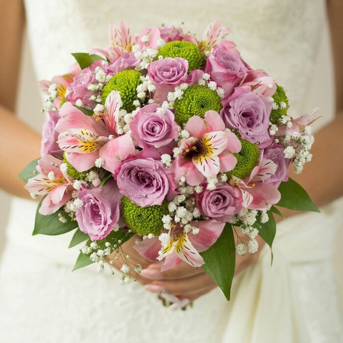 Brimming with charm and artistry, this Epping bridal bouquet is the very essence of romantic celebration, captured in a close-up that draws focus to its lush detail and subtle vibrancy. A profusion of soft pink and lavender roses form the bouquet's spherical heart, some edged in pale blush, their petals layered in rich, inviting spirals-each blossom glowing with the promise of new beginnings. Threaded among the roses are playful pom-pom-like lime green button chrysanthemums, their contemporary brilliance adding joyful contrast and echoing the life found in Epping's well-loved floral parks. Pink alstroemeria, with their trumpet-shaped petals and intricate dark markings, evoke exotic elegance and weave a note of grace and intrigue into the design. Delicate white sprays of baby's breath, soft as early morning mist, fill the spaces between larger blooms, gifting the bouquet its signature airiness and fullness. The subtle gleam of a ring and the gentle curve of the bride's hands around the bound stems suggest quiet happiness and anticipation-accentuated by the creamy, lace-textured background of her gown. Deep green foliage emerges between blossoms, a nod to Epping's leafy avenues and timeless natural beauty. Locally crafted by skilled florists in Epping, this bouquet beautifully blends tradition with fresh modern accents, making it a perfect visual reference for wedding inspiration, local event styling, or stories celebrating the enduring allure of spring and summer nuptials in Essex.