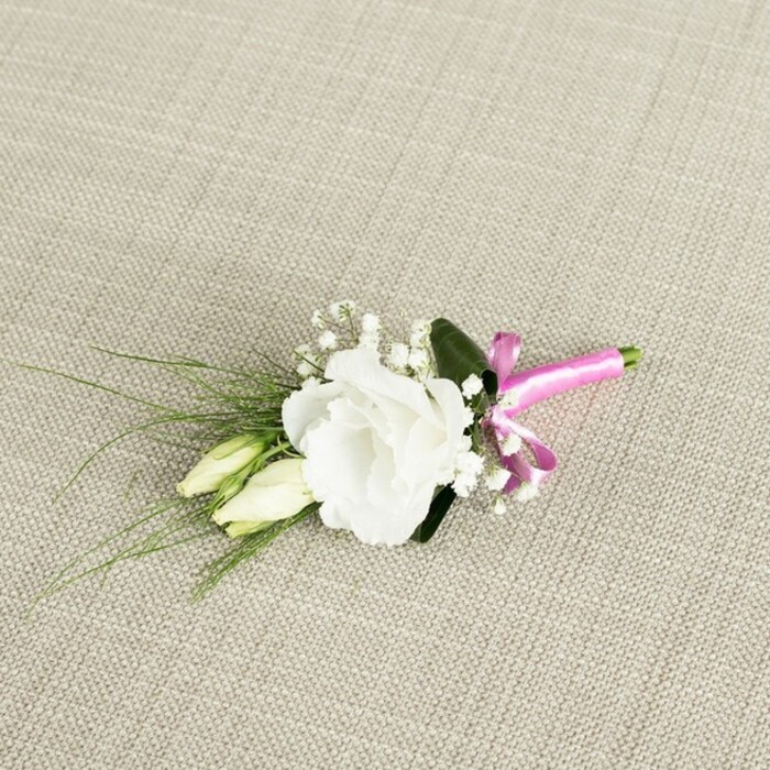 An intricately designed boutonnière is displayed diagonally across a warm, light beige background that resembles finely woven linen, the subtle texture giving the image a soft, organic feel reminiscent of rustic-chic weddings held around Epping's countryside venues. At the heart of the arrangement sits a fully open white lisianthus flower, its many ruffled petals curling outward in layered waves, creating a plush, rounded bloom with a serene, luminous quality. Slightly below and to the left, two elongated buds from the same lisianthus stem nestle closely together, their creamy white and pale green tones gently blending into each other, hinting at blossoms that will open later in the day, just as a celebration unfolds from vows to evening toasts. Threaded delicately throughout the piece are tiny clusters of white gypsophila, their miniature blooms forming a misty halo around the central flower, adding both volume and a sense of lightness, like soft clouds drifting above the trees near Epping Forest. Slender, grass-like strands of greenery sweep out towards the lower left corner, lending dynamic lines and contrast, while a broader, darker green leaf sits discreetly behind the lisianthus, giving the composition depth and a touch of formality. At the base of the neatly trimmed stems, a bold fuchsia satin ribbon wraps tightly around the binding point and is tied into a small but striking bow, its silky surface shining softly against the matte fabric beneath. This bright pink accent provides a modern, joyful twist to the traditional white-and-green wedding palette. The boutonnière feels handcrafted and thoughtful, the kind of floral detail a local Epping couple might choose for a refined ceremony, whether held in town or at a nearby country house, adding a quiet note of elegance to a lapel or dress strap.