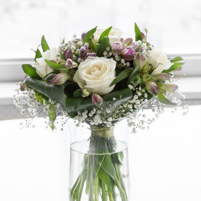 A refined bouquet of creamy white roses, lavender alstroemeria, and clouds of baby's breath is arranged in a clear, cylindrical glass vase, illuminated by soft natural light that feels like a bright yet tranquil morning in Epping. The roses appear in full, luxurious bloom, their petals layered in concentric whorls that create a plush, romantic centre to the design. Nestled among them, alstroemeria in muted lilac and soft purple shades stretch their slender petals outward, some blossoms fully open to show intricate streaks of deeper colour, others still partially wrapped in green sepals that hint at their continuing life. Wisps of white gypsophila weave delicately through the composition, giving the impression of tiny stars scattered across a twilight sky and softening the transitions between the larger blooms. Around and beneath these flowers, glossy green foliage adds depth and dimension, the leaves forming gentle arcs and subtle shadows that lend the arrangement a sense of natural movement. Through the transparent vase you can see the stems gathered carefully and trimmed at an even angle, immersed in clear water that reflects the care and precision typical of an experienced local florist from Flowers Epping. The background is bright and understated, likely a pale wall or sunlit window, keeping the focus entirely on the interplay of whites, purples, and greens. The bouquet's overall feel is serene and uplifting, suitable for a thoughtful sympathy tribute to a family in Theydon Bois, a congratulatory gift for a new home near Epping High Street, or a simple yet sophisticated centrepiece for a Sunday lunch overlooking the edge of Epping Forest. Its balanced design and gentle fragrance evoke calm, affection, and enduring grace.