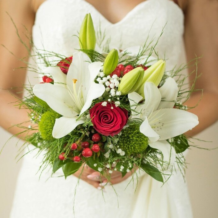 A detailed close-up presents a bride in Epping, her hands gently encircling an exuberant bridal bouquet, framed by the elegant sweep of her white strapless wedding gown. The gown's textured fabric catches soft natural light, accentuating the intricate pattern. The bouquet itself glows with traditional sophistication, led by lush white lilies-some wide open with distinctive gold dust stamens, others tightly sealed in pale green buds waiting to bloom. Nestled among these, a bold, deep red rose draws the gaze, symbolizing unwavering love and making a striking contrast to the serene white flowers. Surrounding the main blooms are fluffy sprays of white baby's breath, small green orbs resembling trick dianthus, and clusters of bright red berries that lend both colour and joyful energy. Tangled in the arrangement, feathery greens-perhaps evocative of Epping's natural woodland borders-and a variety of shiny broad leaves provide fullness and a sense of abundance. The bride's tender grasp at the base of the bouquet seems to ground the arrangement in the present, an emblem of the celebration taking place near the charming woodlands and historic streets of Epping. It's an arrangement that speaks of careful craftsmanship and local tradition-ideal for those envisioning a classic yet vibrant wedding, with florals inspired by the natural palette of their community.