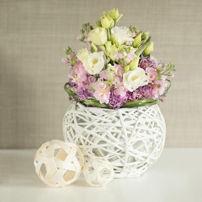 The image presents a meticulously styled floral arrangement that looks perfectly at home in a light-filled Epping dining room, poised on a pristine white tabletop that reflects every gentle hue. At its heart is a distinctive, spherical vase in pure white, fashioned from interwoven strips of rattan or painted branches, forming a delicate open lattice that gives the impression of a handcrafted sculpture. From this airy vessel, a lush bouquet spills outward and upward, dominated by eustoma blooms in creamy white and soft, buttery yellow. Their ruffled, layered petals curl like fine fabric, each flower at a slightly different stage of opening, creating a sense of natural movement and freshness you might associate with early morning walks towards Stonards Hill. Interspersed through the lisianthus are stems of stock in pale pink and dusty lavender, standing a touch taller and adding vertical structure, while their densely packed florets lend a soft, cloud-like texture. At the base of the arrangement, deeper purple filler flowers gather into a rich collar, their smaller, starry heads offering contrast against the larger, smoother blooms above. Encircling this are bands of glossy green foliage, tidy yet relaxed, giving the design a finished, professional feel. To the left on the same white surface sit two decorative spheres crafted from pale, natural wood or rattan, their open basket-weave patterns complementing the vase and reinforcing a theme of organic, modern elegance. The entire scene is framed by a warm beige or greige background with a subtle vertical grain, perhaps a textured wallpaper, which adds depth without distraction. The gentle light, muted colours, and tactile contrasts evoke a sense of calm sophistication, making this arrangement ideal as a spring centrepiece for a family gathering or a refined gift delivered to homes around Epping and the nearby villages.