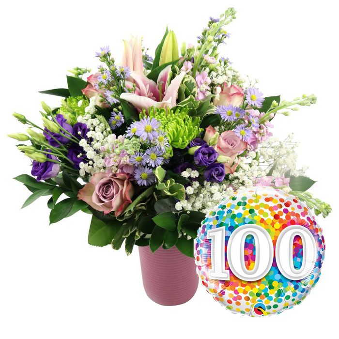 A striking 100th-celebration floral display stands out crisply against a pure white background, evoking the feeling of a carefully prepared gift waiting to be delivered to a home on the edge of Epping Forest. Dominating the left side of the scene is a lush, rounded bouquet, overflowing from a ribbed, matte pink ceramic vase that brings a soft, contemporary touch. The colour palette is an elegant blend of pink, lavender, purple, lime green, and white, thoughtfully balanced to look both festive and refined. Several prominent light pink Oriental lilies rise from the centre and upper tiers of the arrangement, their broad star-shaped petals arching gracefully, while a few closed or half-open buds add height and a sense of unfolding time. Tucked between the lilies, soft pink roses and deeper mauve or dusty rose roses offer classic romance, their petals curled in tight spirals that seem almost velvety to the eye. Interlaced through the roses are deep purple lisianthus blooms, whose smooth, ruffled petals add a luxurious, almost satin-like texture. Small sprays of lavender asters, each with golden-yellow centres, bring a delicate daisy-like charm, reminiscent of wildflowers along lanes leading into Epping. Bright lime green button chrysanthemums dot the bouquet like tiny lanterns, providing refreshing contrast and a modern twist. Airy white baby's breath trails through the design, lifting the overall composition and softening the transitions between each bloom. Surrounding and supporting the flowers, an assortment of rich green foliage adds depth, creating a full, garden-inspired silhouette. To the lower right of the arrangement, slightly overlapping the blooms, a shiny round foil balloon glistens, its surface densely scattered with colourful confetti dots in vivid red, orange, yellow, green, blue, and purple. In the centre of the balloon, large white numerals 100 stand bold and clear with a faint shadowed edge, unmistakably signifying a centennial milestone. The overall impression is one of joy, gratitude, and respect-ideal for marking a 100th birthday, a centenary wedding anniversary, or a long-standing community achievement in Epping with warmth and style.
