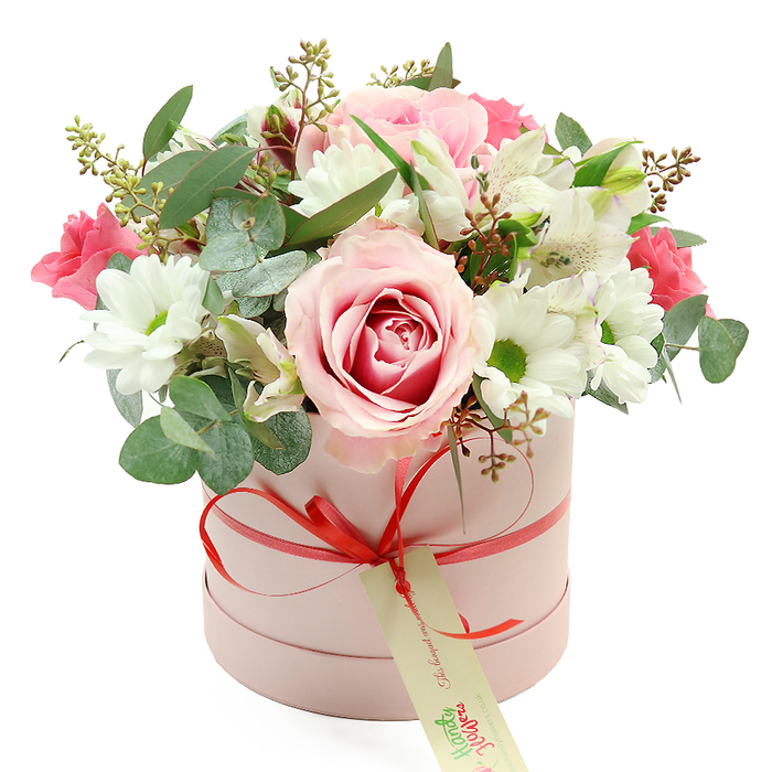 A refined, contemporary hatbox bouquet stands out clearly against a pristine white backdrop, its colours soft yet expressive, like a gentle celebration unfolding in an Epping townhouse. The container is a round, cylindrical box in light pastel pink, its smooth matte surface giving it a velvety appearance. Around the lower edge, a narrow coral-red satin ribbon is wrapped and tied into a compact bow at the front, from which a rectangular cream gift tag with neat green lettering hangs lightly, suggesting a carefully prepared floral gift ready for local delivery. From the open top of the hatbox, a generously full dome of flowers rises, brimming with texture and subtle scent. Several pale pink roses, including one striking bi-colour bloom with a deeper pink centre swirling outwards, form the romantic focal point. These are framed by neat, snow-white chrysanthemum flowers with vivid green centres, their crisp petals lending a clean, uplifting brightness. Threaded between the larger blooms are white alstroemeria, their trumpet-like petals marked with fine speckles, adding an elegant, almost painterly detail. Pops of deeper pink from clustered, smaller flowers-likely spray roses or lisianthus-introduce a richer accent that keeps the palette from feeling too delicate. Lush greenery nestles throughout: rounded, silver-green eucalyptus leaves, darker glossy foliage, and fine stems tipped with tiny buds and seed-like forms, echoing the natural textures of nearby Epping Forest. The overall shape is rounded and balanced, with flowers just spilling over the rim in a way that feels effortlessly abundant. This floral hatbox, reminiscent of boutique designs seen in Epping's more stylish homes and cafes, radiates warmth and thoughtfulness, ideal for birthdays, anniversaries, Mother's Day, or any occasion where you want a gift that feels both modern and heartfelt.