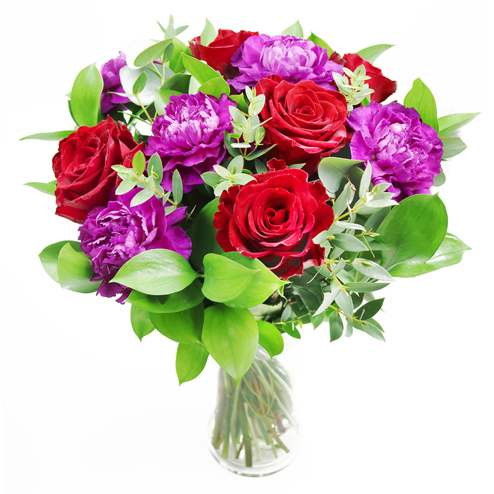 In this arrangement, classic romance and modern style come together in a way that feels perfectly suited to a chic Epping home overlooking the gentle bustle of the High Street. The eye is first drawn to the velvety red roses, each bloom fully unfurled with deep, layered petals that almost seem to glow from within. Their intense colour suggests warmth and devotion, a timeless symbol of love. Intertwined with them are generous purple peonies, their blossoms large and softly rounded, with countless ruffled petals that create subtle shadows and highlights as the light passes over them. The combination of the two blooms forms a rich, jewel-toned palette that feels bold yet carefully considered. Surrounding this floral heart is a textured halo of greenery, where lush, glossy leaves spread outward, creating a sense of abundance. Slender, arching eucalyptus stems add a modern, airy touch, their silvery-green leaves catching the light in a cool counterpoint to the richness of the flowers. The bouquet is cradled in a clear glass vase that reveals the stems beneath, neatly gathered and arranged to echo the care taken above the waterline. Reflections dance in the glass, giving the impression of gentle movement, like dappled light near the edges of Epping Forest on a bright afternoon. Imagined fragrance drifts from the scene: the unmistakable softness of rose, the green freshness of foliage, the faint herbal note of eucalyptus. This bouquet feels ideal for anniversaries, intimate celebrations in homes near Theydon Grove, or as a statement of deep admiration when delivered to someone special in Epping, blending tactile luxury with a clean, contemporary silhouette.