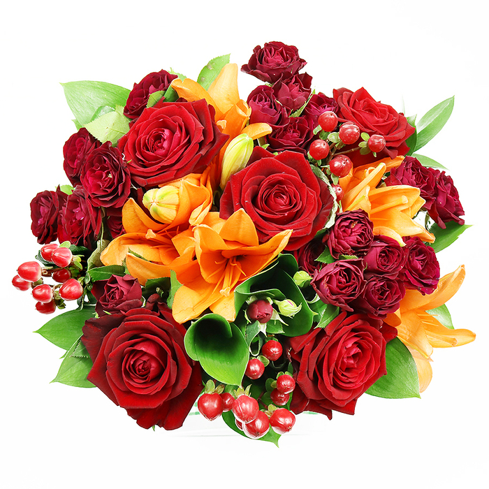 Viewed from a top-down perspective against an immaculate white background, this sumptuous, circular bouquet presents a rich choreography of reds, oranges and verdant green. Several full, deep-red roses anchor the arrangement; their velveteen petals unfurl in concentric waves, offering a tactile, romantic focal point that suggests classic devotion. Orange Asiatic lilies weave through the roses, their bright, trumpet-shaped petals and visible stamens introducing energetic, upward lines - some flowers are boldly open while others hold the promise of bloom in suavely closed buds. Compact clusters of dark red spray roses contribute a dense, brooding depth and a tactile counterpoint to the larger blooms, while lustrous hypericum berries in ruby tones are dotted throughout like tiny, glossy jewels that add both shine and textural contrast. Interspersed ruscus or salal leaves provide generous ribbons of cool green, their glossy surfaces and rounded shapes framing the bouquet and enhancing the visual warmth of the florals. The lighting is soft and even, chosen to preserve colour fidelity and reveal the intricate textures of each petal without creating dramatic shadows. I imagine this design arriving at a home on St. John's Road or being placed on a breakfast table after collection from a nearby florist in Epping; its scent would carry a subtle lift of lily spice balanced by the sweeter, familiar perfume of roses. This arrangement feels celebratory and intimate - a confident gift for anniversaries, autumn weddings, or an elegant expression of love and gratitude.