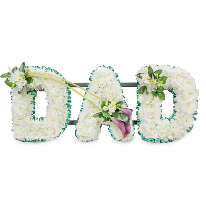 A large funeral flower arrangement spelling out DAD in upright, block letters is captured against a bright white background, the sort of tribute often ordered by families in Epping to honour a beloved father. Each letter is meticulously covered in compact white chrysanthemums, their small, rounded blooms packed so closely that they create a seamless, cushion-like surface. The texture is plush and soft, as though the flowers would yield slightly beneath a fingertip, yet the outlines of each letter remain crisp. Around the outer edges, a vivid emerald green ribbon is carefully gathered and ruffled, running continuously around each letter to provide a strong frame of colour that recalls the green spaces of nearby Epping Forest and the fields surrounding the town. The discreet metal or plastic framework that holds the tribute upright can be glimpsed as thin dark bars behind the letters, hinting at the craftsmanship involved. The first D is adorned at its upper left curve with a compact cluster of white freesia and glossy leaves, adding height and a subtle twist of movement. From near this point, two slender, arching green stems extend diagonally across towards the central A, as if gently sweeping the eye along the tribute. At their meeting point rests an elegant focal spray: two deep purple calla lilies with their smooth, trumpet-shaped petals curl gracefully above a bed of white freesia blooms and fresh greenery. This rich splash of colour offers a dignified contrast to the white base flowers, conveying solemnity and depth of feeling. The final D carries a smaller, harmonising posy of white freesia and foliage on its upper right side, ensuring the design feels balanced from end to end. Overall, the tribute suggests a message of love, remembrance, and quiet strength, a fitting memorial for a father whose presence remains cherished within the local community.
