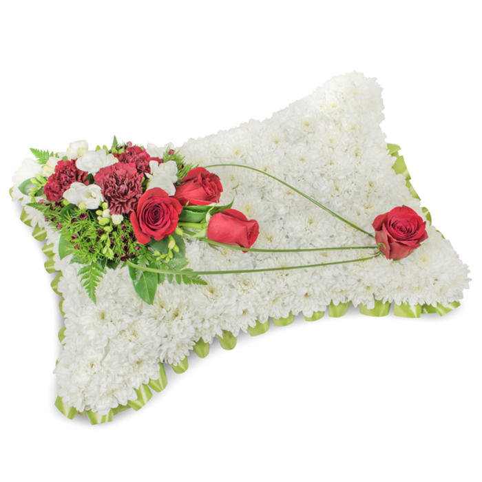 A poignant rectangular memorial cushion crafted with meticulous attention to texture and form rests on a pure white surface. The whole pillow is fashioned from hundreds of small white chrysanthemums, densely assembled so their layered petals form a velvety, soothing field that reads as purity and rest. Along the slightly raised perimeter a whisper of pale green ribbon or delicate foliage peeks out, framing the composition with a soft, fresh outline. Anchoring the design on the upper-left corner is an abundant spray of deep red flowers: several blood-red roses, some unfurling fully, others held in tight bud, paired with deep burgundy carnations that add sculptural, ruffled detail. Scattered among them are tiny ivory blossoms - likely freesias or small spray chrysanthemums - that introduce gentle points of light. Lush greenery, including slender fern fronds and broader glossy leaves, lifts the cluster and gives it a living backdrop. From this focal point three graceful arcs of bright green bear-grass or similar linear foliage stretch diagonally across the white chrysanthemum surface toward the lower-right, imparting movement and a sense of thoughtful connection. At the end of these flowing lines rests a single, flawless red rose, positioned as a quiet emblem of enduring devotion. The overall effect is calm and consoling with a faint, clean floral scent; the pillow is an elegant, traditional tribute suited to services held in local churches or at memorials near Epping Forest and along Station Road. The arrangement speaks of respect, remembrance, and tender craftsmanship.