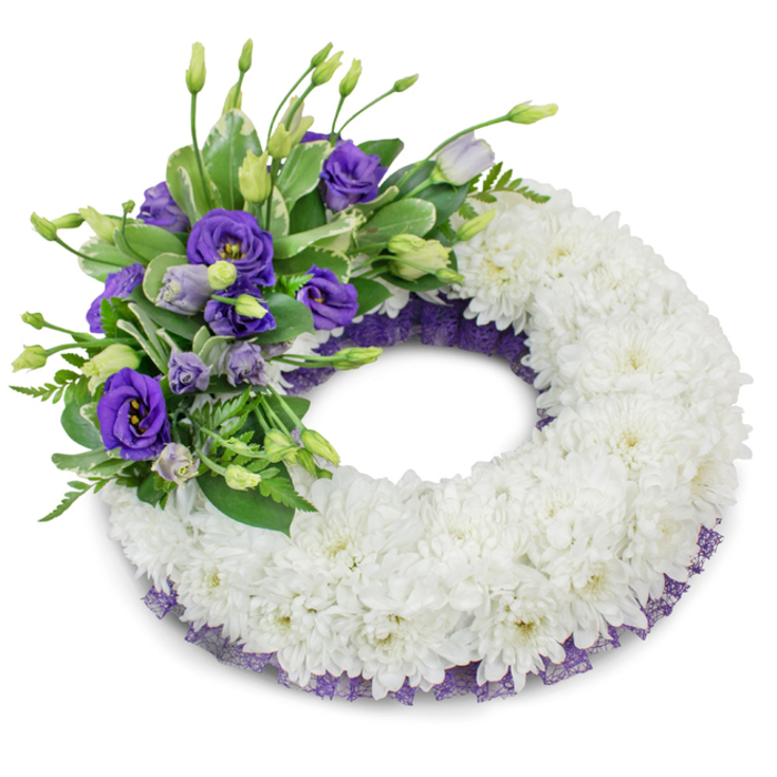 A beautifully arranged sympathy wreath photographed against a pure white background, the kind of memorial piece chosen to express reverence and lasting remembrance. The core of the design is a thick, continuous ring of immaculate white chrysanthemum flowers, their densely packed, ruffled petals creating a soft, pillow-like texture that feels both comforting and dignified. Anchoring the upper-left section is a rich accent cluster of purple Eustoma (lisianthus), the blooms ranging from deep violet opened flowers with visible pale centres to paler lavender buds on delicate stems; this gradient of purple gives the arrangement depth and a gentle rhythm. Interwoven with the lisianthus are varied green foliage elements - glossy broad leaves, some with lighter variegation, and finer, feathery fronds - offering a natural counterpoint of shape, tone and freshness. A whisper of crinkled dark purple mesh or lace sits just beneath the chrysanthemum edge, visible along the wreath's inner and outer circumference and acting as a subtle, elegant trim that ties the palette together. A soft, diffused shadow beneath the wreath on the white surface gives the piece presence and dimension in the photograph, while the imagined scent is mild and green: the clean perfume of fresh chrysanthemums and leafy stems rather than anything overpowering. As a local florist's composition intended for reflection and consolation, this wreath would be fitting for services across Epping - from small chapels near St John's Road to memorials beside Epping Forest paths.