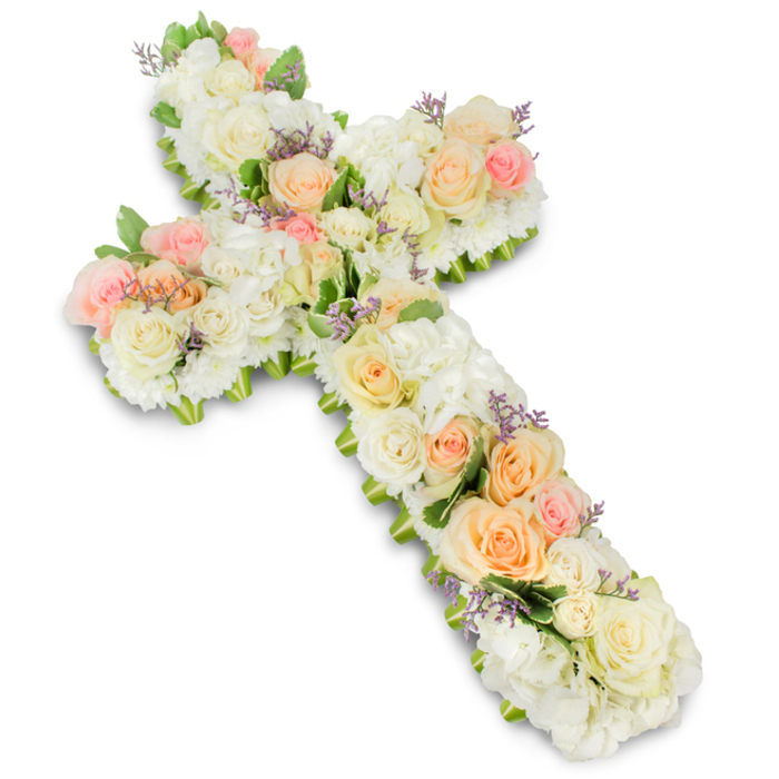 A refined and reverent floral cross dominates the image, positioned centrally against a bright, uncluttered white background that allows every petal and hue to stand out clearly. The cross itself is thick and substantial, composed of an intricate blend of premium blossoms arranged so closely together that they form a continuous, velvety surface. Along the central line and across the arms, clusters of soft peach, apricot, and blush pink roses create gentle focal points, their spiral centers unfolding in delicate layers. Between these roses, creamy ivory and pure white roses mingle with dense heads of white hydrangea or similar rounded blooms, producing a rich tapestry of textures that feels both luxurious and deeply soothing. Tiny touches of pale lavender or light purple filler flowers are interspersed, offering a refined contrast that enhances the pastel tones without disrupting the calm. At the very edges, discreet fronds of fresh green foliage appear, just visible beneath the blossoms, tracing the outline of the Christian cross and giving the piece a natural, finished frame. Subtle, even lighting casts a faint shadow beneath the arrangement, emphasizing its three-dimensional form as though it were resting on a table in a quiet chapel in Epping, perhaps near the historic streets leading towards Epping Forest. The colour palette of whites, creams, pinks, and peaches conveys serenity and compassion, making this piece especially suited for funeral services, chapel viewings, or memorial tributes. Every carefully placed flower reflects the attentive skill of a local florist, expressing sympathy, faith, and lasting remembrance without the need for words.