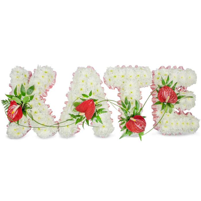 A graceful, bespoke floral tribute spells the name KATE in substantial, capital letterforms, each letter formed from a continuous, dense mat of small white spray chrysanthemums that create a soft, textured surface reminiscent of fine white embroidery. The white field is rimmed with a soft, pleated light-pink ribbon that frames each character with a whisper of colour, enhancing the silhouette and lending a quietly feminine touch. Strategically placed clusters of red anthuriums - their glossy, heart-shaped spathes and central spadices prominent - provide bold focal points that contrast with the serene white foundation. Around and between the letters, lush green foliage including broad glossy leaves, fern fronds and slender trailing vines has been layered to introduce depth, shadow and a touch of wildness that keeps the composition feeling natural rather than purely formal.
Set against a clean white ground with a subtle drop shadow beneath, the arrangement reads with three-dimensional poise, intended as a sympathetic floral tribute for a memorial or funeral setting. One can almost sense the cool, leafy fragrance and the faint powder of chrysanthemum petals; the tactile juxtaposition of soft sprays, glossy anthurium surfaces and ruffled ribbon makes the piece approachable and comforting. As a florist accustomed to commissions across Epping and the surrounding parishes - whether for a town-centre chapel or a service near St John's Road and the edges of Epping Forest - I would recommend this design for families wanting a personalised, dignified expression of remembrance that carries both warmth and restraint.