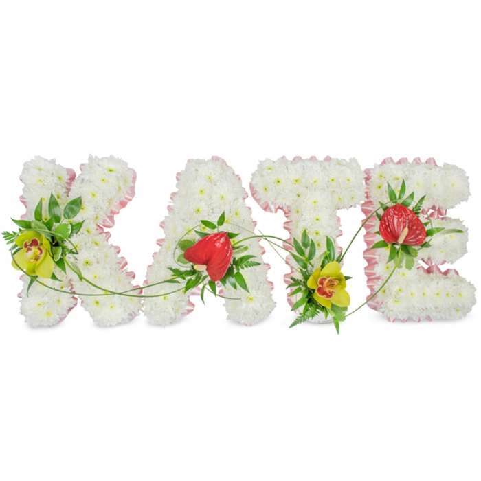 The image shows a refined funeral flower tribute spelling the name KATE in large, crisp capital letters, placed against a plain white background so every bloom and ribbon detail can be appreciated. Each letter is densely packed with small white chrysanthemums, their petals layered and slightly curved, forming a luxuriously full, textured surface reminiscent of soft fabric or freshly fallen snow. Around the perimeter of each letter runs a narrow band of light pink satin ribbon, carefully pleated to follow the shapes of the K, A, T, and E, giving the arrangement a gentle, affectionate touch that feels fitting for a much-loved woman or girl. At the left side of the K and anchored low on the T, vivid yellow-green cymbidium orchids draw the gaze, their star-shaped petals and speckled reddish-brown throats adding sophistication and a hint of exotic beauty. The A and E each carry a single, striking red anthurium bloom, heart-shaped, glossy, and slightly raised from the cushion of white flowers, symbolising love and remembrance with quiet intensity. Surrounding these statement flowers, rich green foliage-broad leaves, feathery ferns, and smaller accent greenery-creates depth and introduces a natural garden feel, while slender green tendrils loop and weave delicately from one letter to another, visually linking the entire name. The overall mood is serene and respectful, yet very personal, the sort of custom memorial tribute often chosen for funeral services in and around Epping, from local churches to woodland services on the edge of Epping Forest, when a family wishes to honour Kate with flowers that spell out her name in a soft yet striking way.