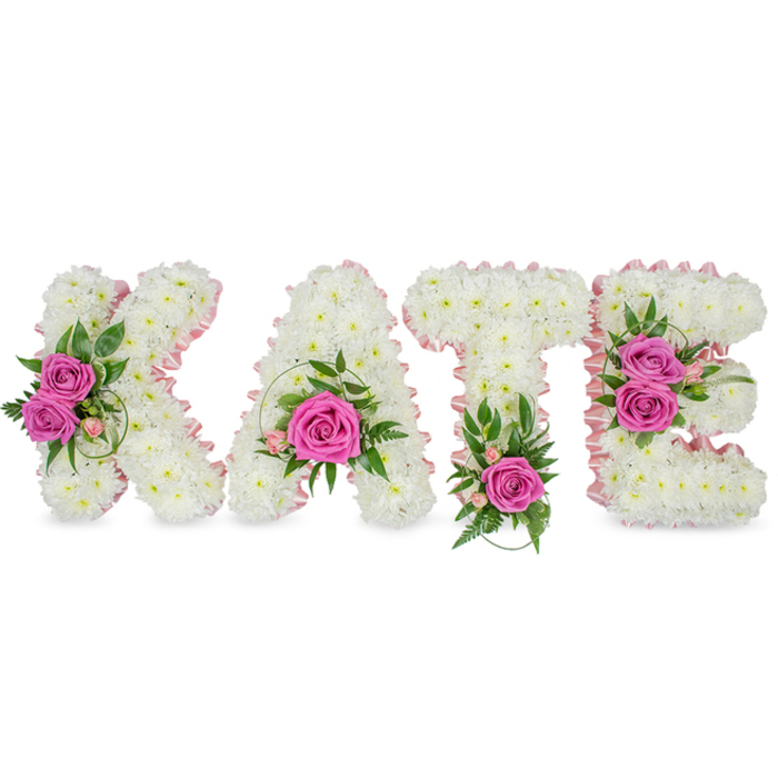 A carefully crafted, three-dimensional floral name tribute spells 'KATE' in bold capital letters, each letter constructed from a dense field of tiny, pristine white chrysanthemums that form a soft, voluminous surface. The inner shapes of the letters are gently emphasized with a pale pink satin ribbon that runs along the contours, offering a refined outline and a soft blush against the snow-white blooms. Each letter is punctuated with clusters of hot-pink roses - their petals unfurling like silk - positioned to draw the eye: the K bears two opened roses to its left, the A cradles a single striking rose near its base, the T is grounded by a solitary vivid rose at its bottom center, and the E carries two roses along its right side. Complementing foliage - variegated leaves, fern-like sprigs and light-pink accent flowers - peeps through the chrysanthemum ground, adding a garden-fresh contrast and delicate texture.
Set against a clean white backdrop with soft shadows that suggest its freestanding form, the arrangement feels luminous in soft morning light and quietly intimate in dimmer chapel lighting. The overall scent is a gentle rose perfume mingled with the clean, faint earthiness of fresh greens. This tailored floral name tribute is often chosen for memorial displays or funeral services and suits the calm, respectful settings found near Epping Market, Bell Common or the local chapels - a beautiful and personal expression of memory, love and farewell.