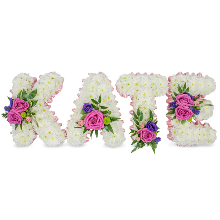 A graceful, custom standing tribute that spells 'KATE' in four three-dimensional letters, presented on a bright white background to make every floral detail visible. Each character - K, A, T and E - is composed of a dense, velvety carpet of pure white chrysanthemums or similar cluster blooms, their many small petals forming a soft, continuous plane that suggests purity and solace. A narrow, pale pink satin ribbon traces the outer and inner edges of each letter, introducing a subtle, romantic blush that gently defines form. Carefully placed clusters of accent flowers break the white with rich colour: prominent deep pink or magenta roses convey love and gratitude, smaller pastel spray roses add tenderness, and darker purple or violet spray chrysanthemums, carnations or lisianthus bring a solemn depth. Framing each cluster is varied greenery-fine fern fronds, smooth eucalyptus leaves and glossy foliage-that adds texture, shadow and a cool, natural scent note. The floral groupings are positioned with considered balance: the 'K' carries a cluster along its diagonal stroke, the 'A' near its lower right curve, the 'T' receives a grounding cluster at its base, and the 'E' shows two accents on its middle arm and bottom section. Bathed in soft, even light, the arrangement reads as a dignified and intimate tribute suitable for funeral services, a celebration of life, or a chapel near Epping Forest or the town High Street. The composition speaks of remembrance, crafted with the quiet expertise of a local florist for families honouring a cherished life.