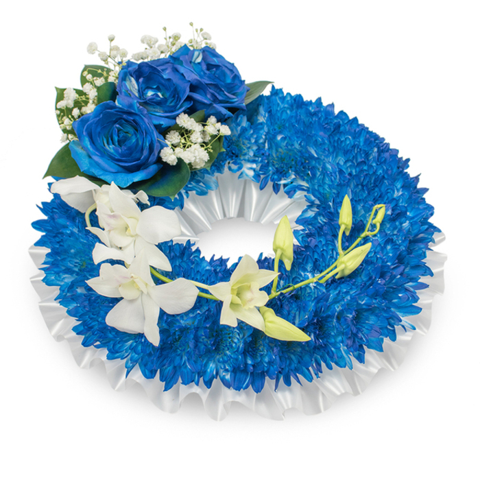 A refined sympathy wreath, crafted in shades of deep blue and bright white and photographed against a stark white background to emphasise its formal silhouette. The principal mass is formed from innumerable royal-blue chrysanthemums, closely arranged to produce a lush, voluminous ring whose fine, spidery petals create a dense, inviting texture. Beneath and lining the inner edge, a broad band of ruffled white satin ribbon sits pleated and soft, catching light with a subtle sheen that articulates the wreath's round geometry. At the upper-left quadrant three large blue roses bloom outward, their layered, velvety petals in a saturated sapphire tone that anchors the arrangement with solemn grace. Accenting these roses are clusters of tiny white baby's breath and a scattering of dusky green leaves that add shadowed depth and a classic condolence touch. From the mid-left the composition is brightened by a cascading run of pristine white orchids, their elongated, silky petals - suggestive of Phalaenopsis-like or Dendrobium blooms - arcing gently across the blue base and guiding the eye toward the lower right. Among the orchids, several fresh green buds stand poised to open, offering a quiet suggestion of continuity. The overall impression is one of tranquil, dignified remembrance: cool blue tones for peace and memory, and pure whites for reverence and solace, a fitting tribute for funeral services in and around Epping, Loughton, or at village halls beyond the forest edge.