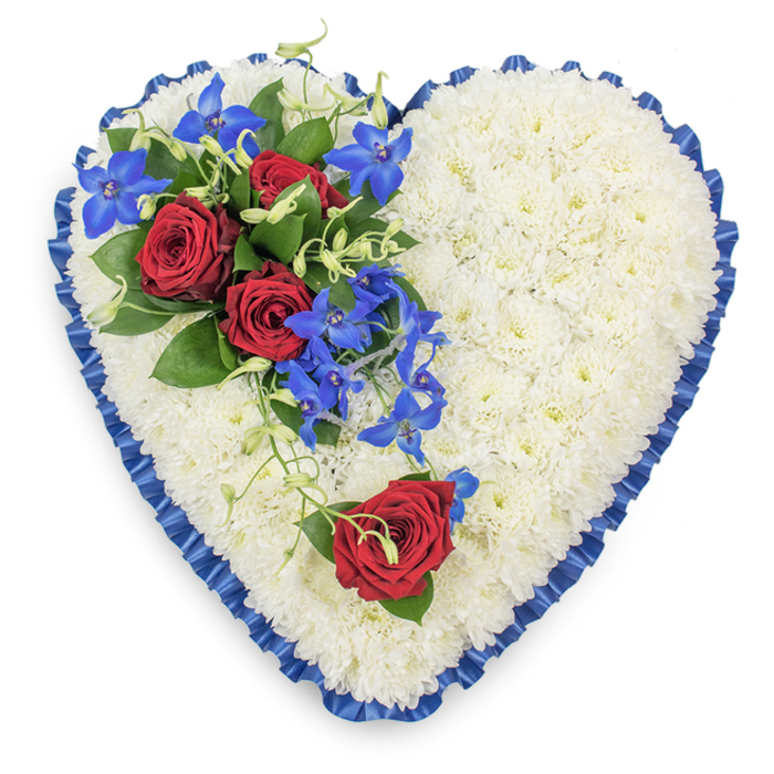 A refined, heart-shaped sympathy tribute dominates the frame, captured from a top-down angle so every petal and ribbon fold can be clearly imagined. The heart itself is completely filled with compact, snow-white chrysanthemums, their small, rounded petals pressed closely together to form a smooth yet delicately textured surface, soft and cushion-like to the eye. This white expanse conveys purity and peace, reminiscent of quiet winter light over Epping's churchyards and tranquil residential streets. Encircling the entire shape is a royal blue satin ribbon, pleated by hand into gentle ruffles that rise and fall around the edges. The fabric reflects the light with a subtle shimmer, creating an elegant border that defines the heart and adds depth to the design. At the upper left, an asymmetric spray of vibrant flowers provides a rich contrast. Here, three deep red roses, fully open and velvety, rest among dark, polished green leaves. Their crimson colour expresses enduring love, loss, and respect. Interlaced with them are bright blue dendrobium orchids, their slender, star-shaped blossoms glowing with electric blue tones that spill gracefully down towards the centre of the heart. Light green curly tendrils thread through the orchids and roses, creating a sense of gentle motion and natural softness. On the lower right curve of the heart, a single, equally vivid red rose is placed as a quiet counterpart to the main cluster, suggesting one special person held dearly in memory. The overall feel of this piece is calm, dignified, and heartfelt-a bespoke funeral tribute that a local Epping florist might prepare to accompany a final goodbye, whether at the crematorium, a village church, or a family gathering of remembrance.