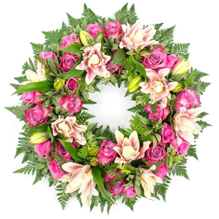 This image shows a large, meticulously crafted circular wreath set against a stark white background, which frames the flowers like a gallery backdrop. The wreath itself is full to the edge, composed of a rich tapestry of pink and cream blooms layered over deep greenery. Dominating the design are roses in saturated fuchsia and magenta tones, mingled with gentler blush pinks. Each rose exhibits a different stage of bloom: some are wide open, revealing intricate ruffled centres, while others are partially unfurled, with outer petals loosely cupping the heart of the flower. Nestled among these roses are luxuriant double lilies in pale pink, creamy ivory, and hints of peach, their petals slightly curled and often edged with a deeper tone, giving a subtle starburst effect. Here and there, lime-green lily buds rise slightly above the main flowers, adding height and a sense of fresh growth. The entire arrangement is underpinned by a cushion of foliage in multiple textures. Fine, dark fern fronds trace a soft fringe around the ring, while larger, glossy leaves and occasional lighter green, almost variegated accents fill spaces between the blooms, ensuring no gaps and providing a natural, woodland feel. The lighting falls evenly across the wreath, emphasising the velvety texture of the roses and the satiny finish of the lilies, as if the tribute had just been prepared in a quiet corner of an Epping flower shop ready for delivery to a local chapel or cemetery. The overall effect is elegant, serene, and deeply respectful, suitable for funeral services, memorials, or commemorations where loved ones in Epping wish to express sympathy and remembrance with enduring floral beauty.