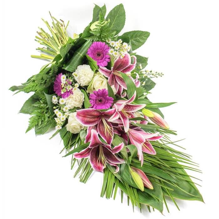 A beautifully composed funeral sheaf in shades of fuchsia, white, and deep green is shown against a bright white background, every element sharply defined as though laid out on a chapel table in Epping before being taken to a service. The arrangement stretches diagonally from the securely bound stems at the upper left to a gentle, tapered point at the bottom right, forming a classic teardrop silhouette associated with sympathy and remembrance. Dominating the centre, several large Stargazer lilies unfold their richly coloured petals, each bloom a star of vivid magenta-pink with snowy white edging and fine speckles in burgundy tones. Some lilies are fully open, their stamens visible, while others remain as plump buds tinged with pink and green, adding a sense of unfolding time and lingering presence. Interwoven with the lilies are bold magenta gerbera daisies, their round, daisy-like heads offering an intense pop of colour that feels modern yet tender. Around them, clusters of pristine white roses and softly frilled white carnations provide a gentle contrast, their creamy shades symbolising reverence, purity, and quiet love. Tiny white filler blooms, reminiscent of daisies, tuck into the gaps, giving lightness and intricacy to the design. A rich surround of foliage frames the flowers: broad, glossy dark green leaves form the base structure, while airy fern fronds and fine, grass-like blades spill gracefully outward and downward, emphasising the spray's length. The lighting is clean and even, casting no harsh shadows and preserving a calm, respectful mood. This thoughtfully balanced tribute, similar to those often chosen for funerals and memorials across Epping and nearby villages, offers a dignified way to honour someone cherished and deeply missed.