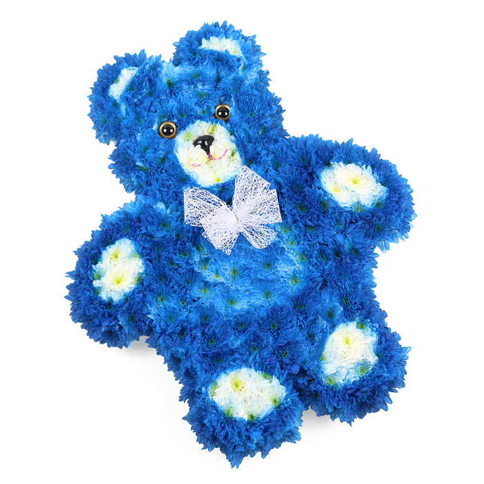 This image shows a lovingly crafted floral teddy bear tribute resting on a pure white background, the kind of intricate design often chosen by families in Epping when words feel too small. The bear itself is shaped with precision: a rounded head, plump torso, and short, softly curved arms and legs, all built from a dense carpet of royal blue flowers. These tiny, uniform blooms, likely chrysanthemums or a similar spray variety, are packed so closely that they create a smooth, plush-like surface, echoing the familiar hug of a favourite childhood toy. Soft cream to pale yellow flowers are thoughtfully placed to form the inner ears, the round muzzle, and four neat, oval paw pads at the tips of each limb, providing a gentle contrast that outlines the bear's features without disturbing the soothing blue palette. Dark button-style eyes and a small triangular nose, both in deep, glossy tones, add character and warmth, while a slender curved line beneath the nose suggests a subtle, comforting smile. Around the bear's neck, a sheer white mesh bow is tied, its translucent loops and tails giving a light, airy finish that hints at innocence and purity. Photographed from above, the arrangement lies flat, tilted just enough to feel natural and serene, with the crisp lighting highlighting every petal and contour. This floral teddy bear is often chosen as a heartfelt funeral or memorial tribute for a baby boy or young child in the Epping area, bringing a gentle presence of love and remembrance to services at local churches, crematoriums, or quiet family gatherings at home.