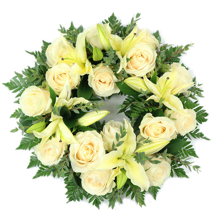 A large, circular funeral wreath shown flat on a white background, designed to embody calm and respectful elegance through its restrained palette of cream, pale yellow and deep green. The composition is densely layered: clusters of creamy roses-fifteen to twenty in number-sit prominently, each bloom revealing the classic, concentric coils of rose petals that catch the light and read as soft ivory. Scattered between these are substantial lemon-tinged lilies, their trumpet forms providing vertical emphasis and sculptural grace; some lilies are fully open, exposing slender stamens and the gentle sweep of each petal, while others remain in bud, suggesting quiet promise and variety of texture. Underpinning the flowers is a generous base of foliage-feathery fern fronds that lend a gentle, whispering silhouette and darker, broader-leaf sprigs reminiscent of eucalyptus that introduce deeper green tones and a matte finish. The photographed perspective and soft natural illumination highlight the tactile differences between glossy blooms and the more fibrous greenery, and one can almost sense the subtle fragrance: the roses' delicate powder and the lilies' green-sweet lift. Crafted for memorial use, this wreath's perfect circular shape signifies continuity and remembrance and is appropriate for placement at funerals, chapel services or graveside moments across the Epping area-from the bustle of the High Street to quiet corners near Epping Forest. The arrangement is made with considered skill, offering solace and an understated expression of sympathy.