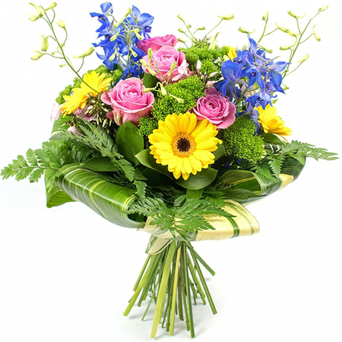 In this image, a professionally arranged, hand-tied bouquet stands proud against a clean white backdrop, every colour and detail clearly visible, as though displayed on the counter of a trusted Epping flower shop. The focal point is a cluster of large pink roses, their petals fully open and curving in soft layers that convey timeless romance and tenderness. Interwoven with these roses are bright, sunshine-yellow gerbera daisies, each bloom displaying a dark brown centre that draws the eye inward and creates a lively contrast against the smooth, pastel rose petals. From within this rounded heart of colour, tall stems of rich blue delphinium reach upward, adding vertical rhythm and a cool, almost airy quality, like glimpses of blue sky above the greenery of nearby Epping Forest. Around the flowers, an abundance of foliage gives the bouquet a generous, garden-inspired look. Delicate fern fronds spill gently from the lower edges, introducing a feathery texture that softens the silhouette. Broader, glossy green leaves sit closer to the blooms, their polished surfaces catching the light and deepening the sense of freshness. Dotted throughout are small clusters of bright, textured filler foliage in lively lime and grassy greens, building depth and structure so that every blossom appears carefully supported. Slender, pale green stems tipped with unopened buds rise here and there, lending a sense of movement and the promise of continued blooming. At the base, several large, arched leaves, some subtly striped in lighter green, are wrapped neatly around the stems to form a natural collar. Beneath this, the stems are gathered and tied with a pale, textured wrap that hints at natural paper or fabric. The overall design feels joyful yet refined, perfectly suited for birthdays, congratulations, or a thank-you bouquet delivered to homes and workplaces across Epping and the surrounding lanes.