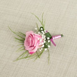 Displayed against a neutral, woven beige linen that looks like a carefully chosen backdrop in a boutique Epping florist studio, this blush pink rose boutonniere captures the quiet romance of a wedding morning. The rose sits at the centre of the image, semi-open and velvety, with petals that transition from a soft, powdery pastel at the heart to a richer, more saturated pink at the curled edges, as if holding the warmth of a late afternoon sun over Epping Forest. Around its base, dainty clusters of white baby's breath create a frothy, cloud-like ring, each tiny flowerhead adding texture and lightness, softening the boldness of the rose while reinforcing its classic charm. Thin, wispy strands of bright green, grass-like foliage radiate gently from behind, giving the design a sense of movement and freshness, reminiscent of the greenery lining Bower Hill and the nearby fields. A single broader, darker green leaf is folded and wrapped close to the stems, forming a neat, structured cradle that grounds the arrangement. The stems are tightly bound in a sleek satin ribbon blending lavender-pink with deeper magenta-purple hues, tied in a discreet knot that suggests refinement rather than showiness. The ribbon's sheen catches the soft, even light, creating subtle highlights that echo the natural gloss of the rose petals. Every element of the boutonniere speaks of considered craftsmanship and balance, the kind of detail a groom or groomsman might wear for a ceremony at an Epping church or a prom at a local venue, adding a small but meaningful flourish to a day centered on love and celebration.