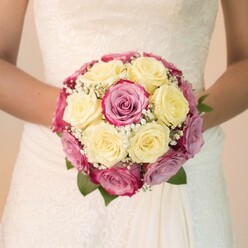 This evocative image offers a close, eye-level perspective, focusing intently on the artistry of a lush, handcrafted bridal bouquet held gently by a bride in one of Epping's timeless wedding venues. The bouquet, meticulously arranged for natural balance and flair, blends creamy lemon-yellow roses with vivid fuchsia and soft mauve-pink roses, whose petals carry gentle hints of lavender-each bloom perfectly selected and arranged by a skilled hand familiar with the quiet elegance of Epping's countryside. Interspersed among the colourful roses, sprigs of white baby's breath weave throughout, suffusing the bouquet with a light, ethereal quality reminiscent of early spring mornings in Stonards Hill parklands. Nearby, deep green leaves peek out, their rich hues reflecting the rural beauty of the local area. Illuminating the flowers, pure daylight highlights the fresh, delicate petal textures and the scalloped contours of fine lacework visible on the bride's bodice. The trailing skirt, made from smooth ivory fabric, cascades beneath the bouquet, its gentle folds hinting at graceful movement just steps from Epping's Market Square. The bride's forearms form a protective circle around the bouquet, suggesting the careful joy and anticipation that mark the opening chapter of any love story. Blurred and light-toned, the background allows the bouquet and gown to stand in striking relief, making this image as much a celebration of local floral talent as it is of romance. Ideal for couples searching for wedding inspiration in Epping, this photograph radiates both sophistication and devotion.