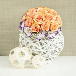 On a smooth white table, a carefully composed floral centerpiece radiates quiet luxury, the sort of arrangement you might find gracing a wedding table at a country venue just outside Epping. The core of the design is a lush, spherical cluster of peach and apricot roses, each head closely nestled to its neighbour so that not a single stem shows. Their petals are full and velvety, curling outwards in soft spirals that create a gentle gradient of warm pastel tones, from soft blush at the tips to deeper apricot towards the centre. Encircling the bottom of this rose dome is a ring of elegant white lisianthus, each bloom edged in a rich violet or royal purple that looks almost ink-drawn. This coloured trim offers a sophisticated contrast to the warm roses, suggesting a sense of occasion without overwhelming the eye. Between the lisianthus, fine sprigs of baby's breath emerge, their tiny white blooms adding a misty lightness, like a soft veil. These flowers sit securely within a rounded white container woven from rattan or wicker into an intricate, open-lattice globe. The container's clean white tone reflects the light and enhances the freshness of the flowers, while its airy pattern adds depth and shadows. Two natural-toned decorative spheres rest in front, larger and smaller, woven from pale wood-like strands that mirror the basket's texture. Behind it all, a beige fabric backdrop with a subtle weave brings a warm, neutral calm, much like the relaxed interiors of homes around Theydon Bois and Coopersale. The composition feels serene, romantic, and meticulously designed, ideal for marking special moments or adding an understated touch of celebration to an Epping dining room.