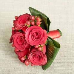 A compact yet luxurious bouquet of red roses and berries is artfully displayed on a pale beige, finely textured fabric, its romantic colours glowing against the calm background. At the centre, several large roses, fully opened, reveal layers of velvety petals in deep crimson, with hints of darker shadow near the heart of each bloom and softer scarlet tones along the outer edges. The roses are fresh and full, their rounded forms snugly gathered so they almost touch, creating a sense of abundance despite the bouquet's modest size. Tucked between them, small clusters of round Hypericum berries in red and rosy orange peek through, their smooth, glossy skins catching the light and adding a playful yet refined contrast to the softness of the petals. Surrounding the flowers, wide, dark green leaves are elegantly curled and folded, forming a supportive green cradle that lifts the bouquet and frames it like a living sculpture. A narrow band of matching red satin ribbon can be seen binding the stems, its polished finish echoing the richness of the blooms. The lighting is gentle and diffused, enhancing the velvety textures and creating a calm mood. This arrangement feels like something a bridesmaid might carry at an intimate wedding in Epping, perhaps near the station or a tucked-away venue off High Street, or like a treasured thank-you gift hand-delivered to a doorstep. Crafted with the precision and sensitivity of an experienced local florist, it speaks of love, heartfelt gratitude, and the quiet thrill of a shared moment.